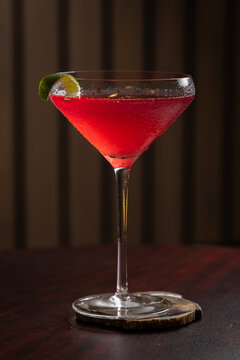 Pink cosmopolitan cocktail in martini coupe glass. Lime twist adorns the rim. Dark wood table create depth. Sleek, elegant, sophisticated mood