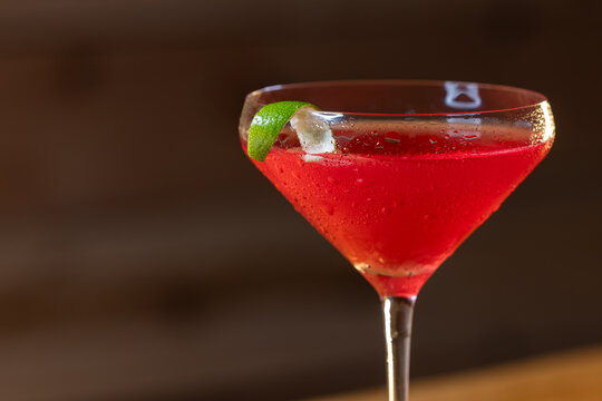 Pink cosmopolitan cocktail in martini coupe glass. Lime twist adorns the rim. Dark wood table create depth. Sleek, elegant, sophisticated mood