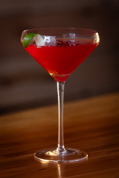 Pink cosmopolitan cocktail in martini coupe glass. Lime twist adorns the rim. Dark wood table create depth. Sleek, elegant, sophisticated mood