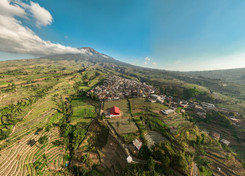 Aerial view of Kruwisan village nestled among terraced agricultural fields with Mount Sindoro in the background under a clear blue sky in Kledung, Central Java, Indonesia.