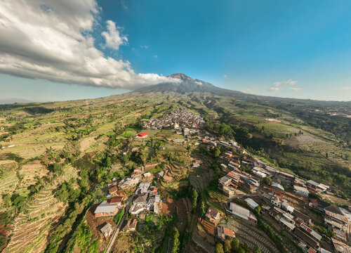 Aerial view of Kruwisan village nestled at the foot of a majestic mountain, surrounded by lush terraced agricultural fields in Kledung, Central Java, Indonesia.