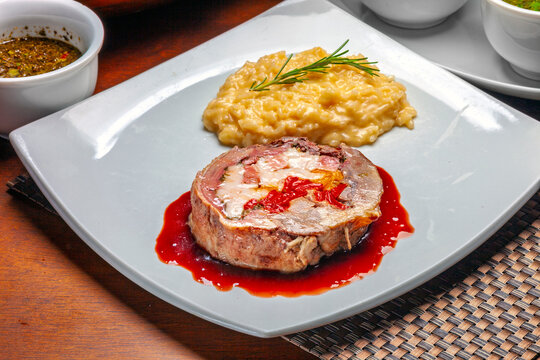 Gourmet stuffed meat roulade served with creamy risotto and rich red sauce on a white square plate.