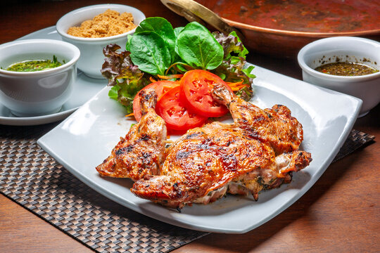 Delicious roasted spring chicken (galeto) on a white plate with fresh salad, farofa, and sauces over a wooden table. Traditional Brazilian food.