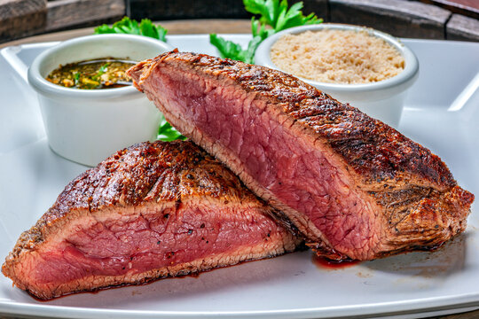 Bife grelhado malpassado e fatiado em um prato branco, com farofa tradicional e molho chimichurri. Churrasco brasileiro.