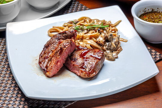 Juicy medium-rare grilled beef steak served with creamy mushroom pasta and chimichurri sauce on a white plate.