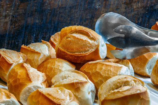 Freshly baked crusty bread rolls in a bakery display with metal tongs picking one up against a rustic wooden background.