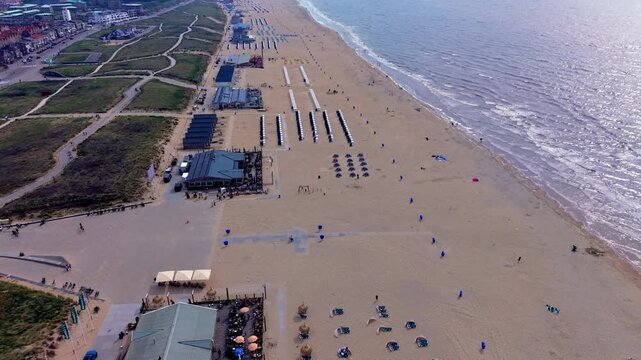 Wide horizontal aerial drone video of Katwijk aan Zee beach clubs, sun loungers, North Sea waves, sun glint reflections, sand dunes, and boulevard in South Holland, Netherlands on afternoon