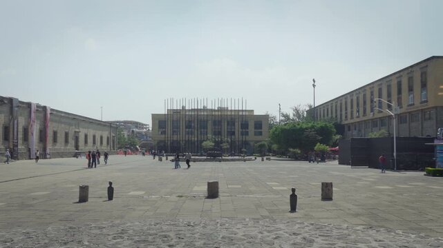 Pedestrians walking across public Plaza Tapatia square featuring central flagpoles with historic building in background and Hospicio Cabanas museum facade on left during bright daylight