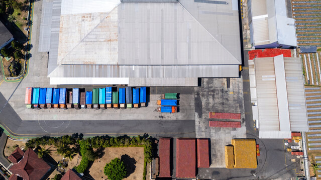 Aerial view of a logistics warehouse facility with parked semi-trucks, colorful shipping containers, industrial buildings, and stacks of materials in Salatiga, Central Java, Indonesia.