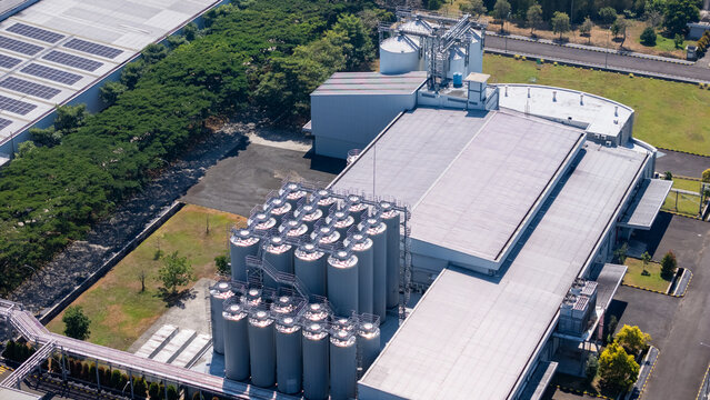Aerial view of an industrial facility featuring a cluster of large metal storage silos, a flat-roofed warehouse building, and surrounding green space in Salatiga, Central Java, Indonesia.