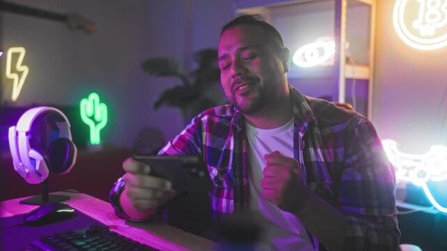 Man holding smartphone and smiling while gaming at studio desk with headset, keyboard and neon cactus sign visible; joyful focus.