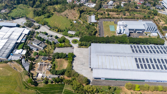 Aerial view of a large industrial factory complex with solar panels on the roof, surrounded by lush green fields and trees in Salatiga, Central Java, Indonesia.