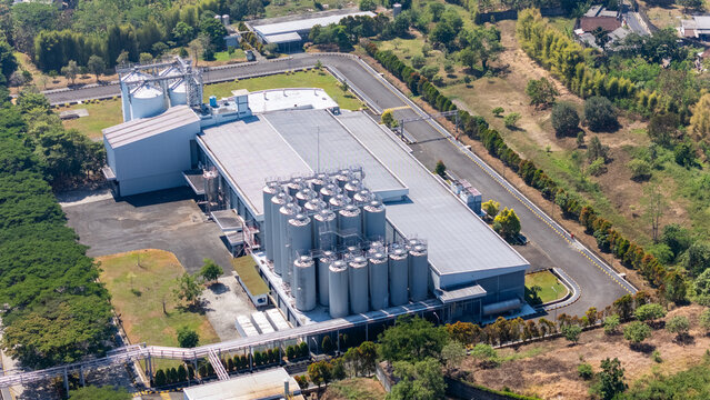 Aerial view of an industrial factory complex with large metal storage silos and a flat-roofed building surrounded by paved roads and greenery in Salatiga, Central Java, Indonesia.