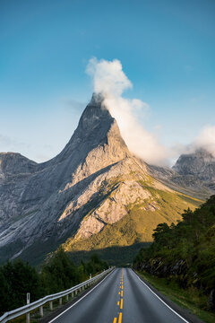 Aerial view of Stetind mountain peak rising above an asphalt road with yellow markings, surrounded by rocky slopes, pine trees, and clouds in Narvik Municipality, Nordland, Norway.