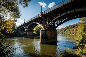 Fototapeta premium Metal truss bridge spans river under blue sky with autumn trees. Stone pillars support structure crossing water. Historic engineering marvel connects landscapes.