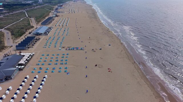 Wide horizontal aerial drone video of Katwijk aan Zee sandy beach, white resort cabins, seaside pavilions, North Sea foam waves, and coastal dunes in South Holland, Netherlands on late afternoon