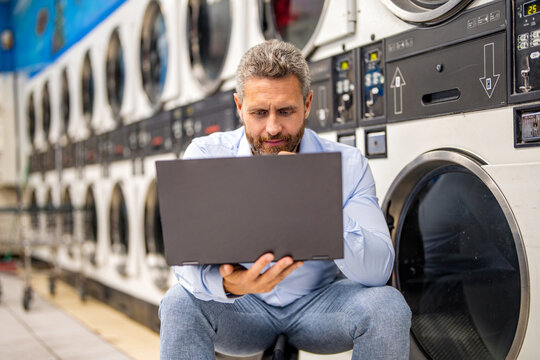 Doing housework. Busy man working on laptop at laundry. Overloaded employee multitasking laundry and job. Concentrated worker dealing laundry and deadlines. Laundry overload for busy businessman