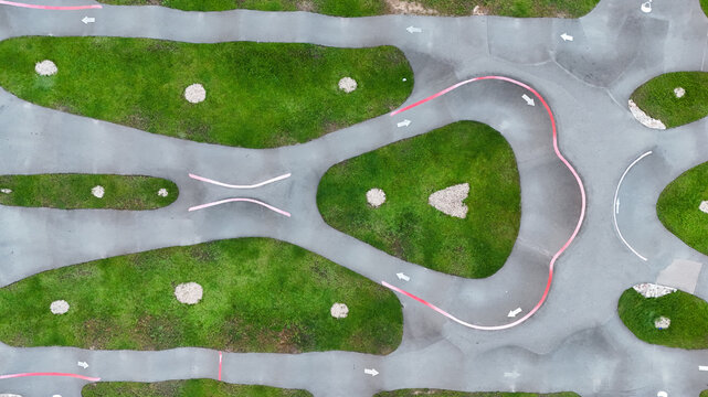 Aerial view of a paved pumptrack with grass patches and directional arrows in a public park in Riga, Latvia.