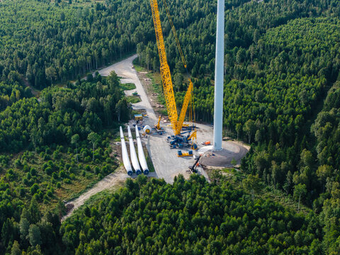 Aerial view of wind turbine construction at Targale Wind Park, with a large yellow crane assembling blades and tower sections amidst a dense forest in Targale, Ventspils Municipality, Latvia.
