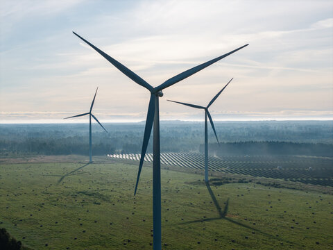 Aerial view of wind turbines standing in a green field with rows of solar panels in the background under a soft morning sky in Targale, Ventspils Municipality, Latvia.