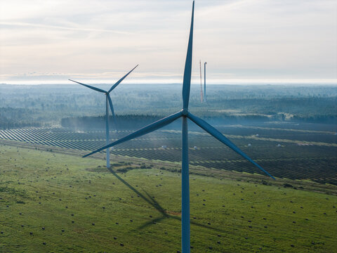 Aerial view of wind turbines standing in a green field with rows of solar panels in the background under a soft morning sky in Targale, Ventspils Municipality, Latvia.