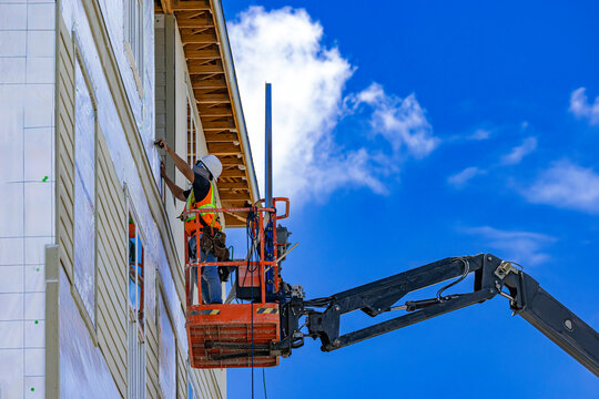 Construction worker installing exterior siding from cherry picker boom lift on building facade in USA