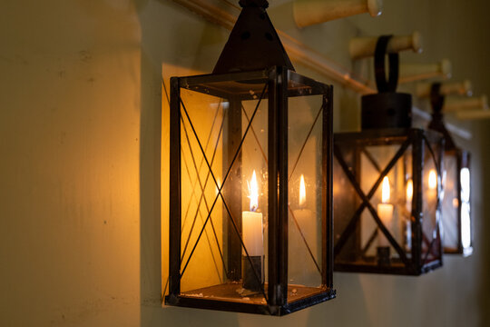 Three vintage glass hurricane lanterns hanging from a wooden pegboard. The interior of the house has wooden walls, bathed in the golden glow of lit tapered candles. The old lanterns are made from bras