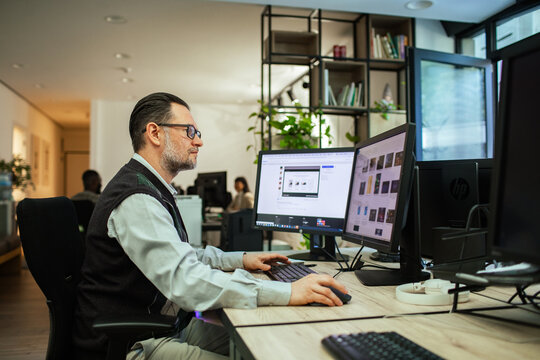 Mature designer working at dual monitors in modern open office