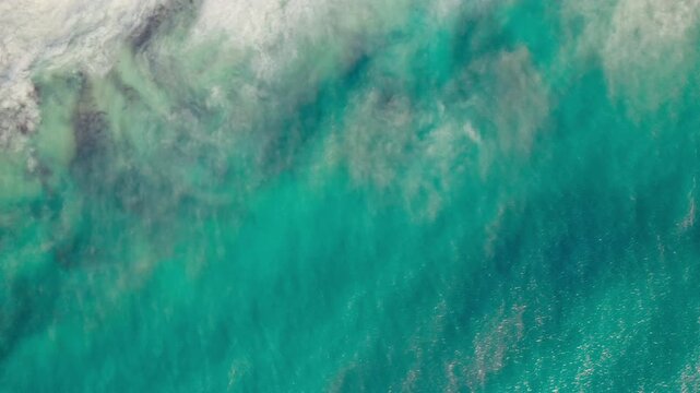 Clear blue water crashes against the shoreline in view from directly overhead during a bright sunny afternoon