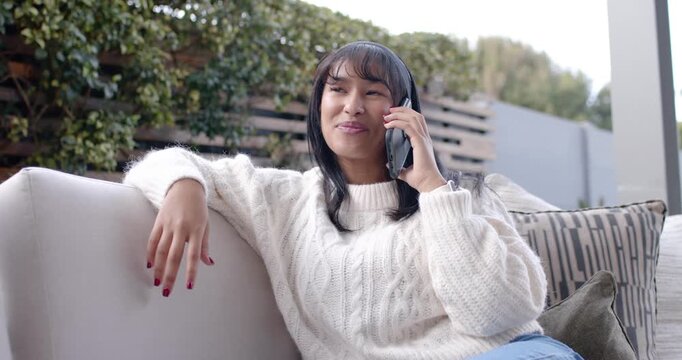Adult Asian woman taking call, wearing cream sweater on sofa, chatting with friend, tapping phone
