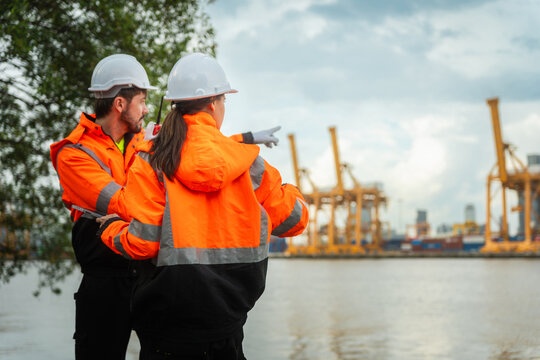 Logistic managers in safety gear standing together and pointing at cargo port terminal, professional industrial engineers team discussing project while pointing forward at shipping port yard
