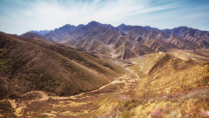 A panoramic view of a rugged, arid mountain valley stretching under a clear blue sky, with sparse vegetation and exposed terrain. © tai