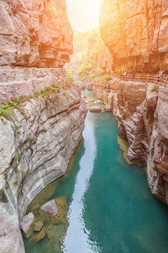 A narrow canyon with turquoise river flows between red rock cliffs, with a wooden path and visitors at sunrise.