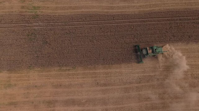 Agricultural machinery operates in a large field, moving steadily to harvest crops, with golden grains and soil visible in the background
