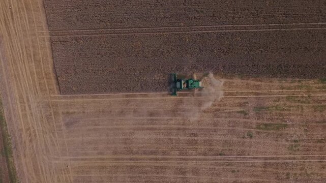 Aerial perspective shows green tractor actively harvesting crops in expansive agricultural field, with dust clouds forming as it travels along the rows