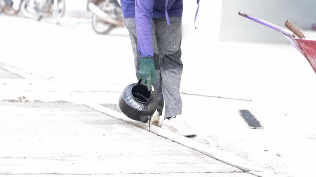 Construction worker pouring liquid black asphalt joint sealant into a concrete expansion gap on a outdoor road surface.