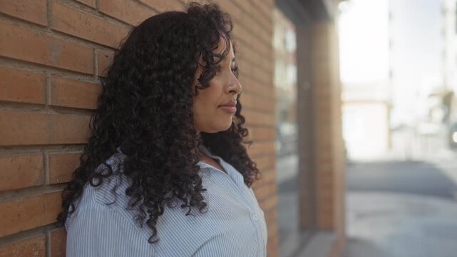 Woman with curly hair leans against brick wall, profile face visible, looking sideways next to a building facade; calm reflection.