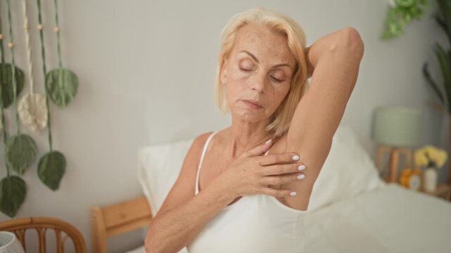 Woman touching armpit and inspecting underarm skin on bed in bedroom, hand placed underarm in skincare routine; self care calm.
