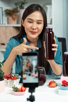 Smiling young beautiful Asian reviewing tasty mixed strawberry and cherry juice detox drinks, showing healthy product to promote for special promotion selling on social media online record. Stratagem.