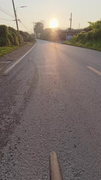 POV road cycling along a quiet rural road at golden hour, with warm sunlight, green vegetation, roadside homes and a peaceful countryside atmosphere.