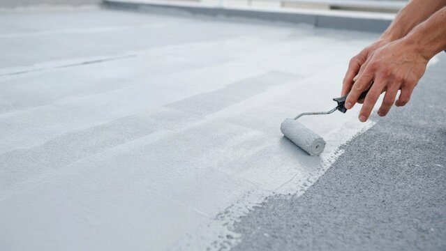 Worker hands applying grey protective liquid waterproofing coating with paint roller on concrete roof surface close up view.