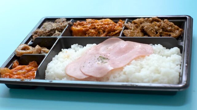 Side view of a woman's hands opening the lid of plastic lunch box containing Korean food, set against a blue background.