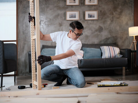 Asian mature man self-assembling wooden flat-pack furniture with DIY carpentry tools, following instruction manual, home improvement, craft installation, hobby workshop, handyman active lifestyle.