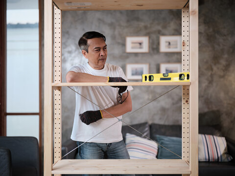 Asian mature man checks his self-assembling wooden furniture with DIY tools, accuracy achievement satisfied, cheerful and proud home improvement accomplishment, hobby carpentry workshop handyman.