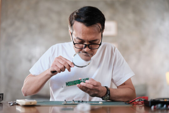 Mature Asian man looks at and considers circuit board details with magnifying glass, focusing on precision checks for home electrical appliance DIY fix, electronics component hobby, handyman workshop.