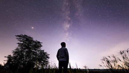 A silhouetted individual stands outdoors at night, gazing up at the starry sky with the Milky Way visible above natural landscape elements. © tai