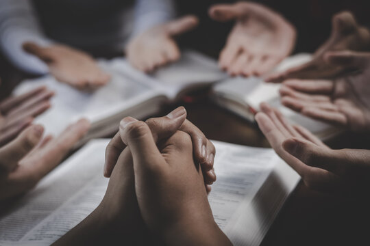A Christian prayer group joins hands around a table with an open Bible and cross, sharing faith, worship, and spiritual connection with God.