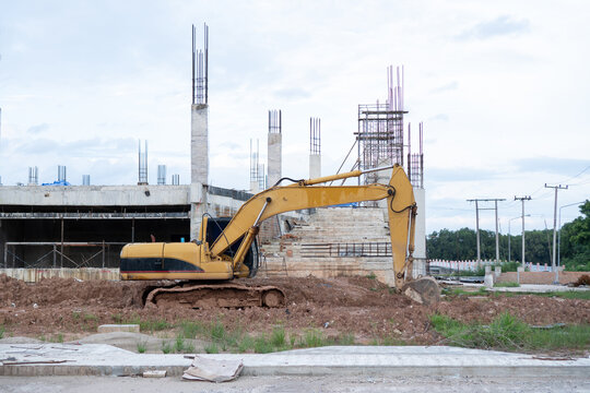 Yellow excavator working on active construction site with heavy machinery, concrete building structures, dirt ground under bright sky, showing powerful industrious mood and focused energy