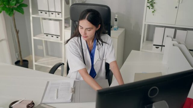 Female doctor typing on computer while reviewing patient record in hospital. 