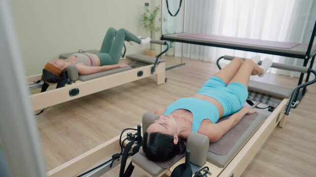 Two young women exercise on Pilates reformer machine in fitness studio. 